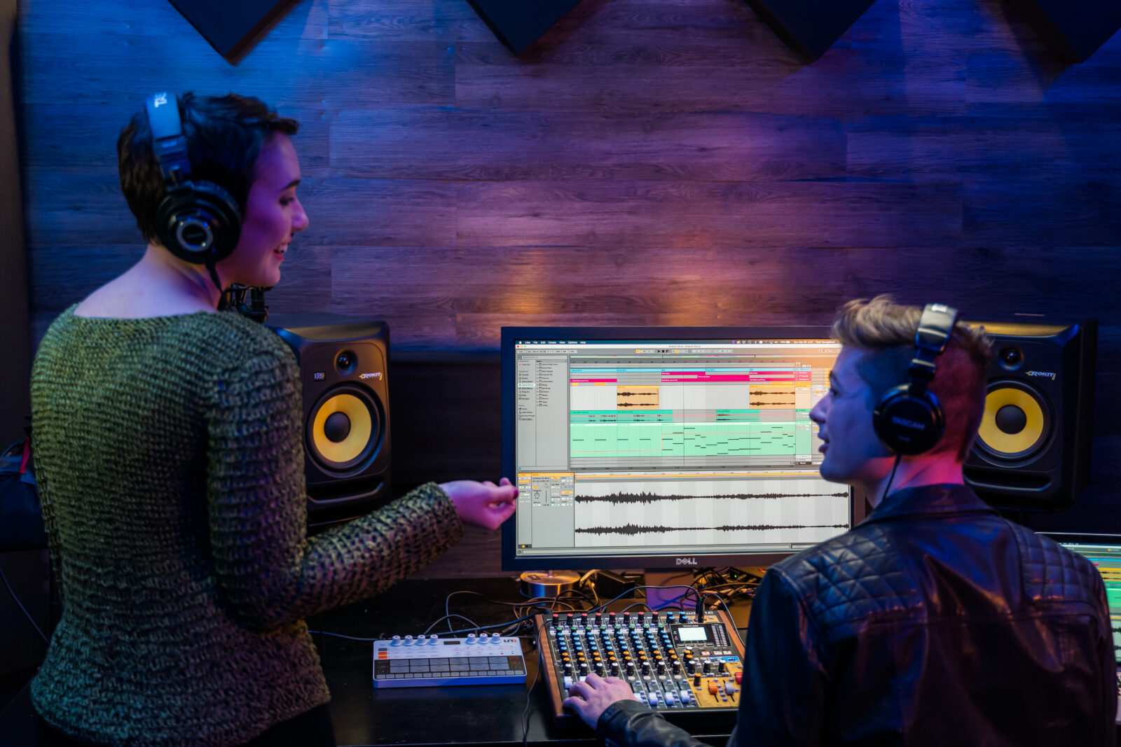 Two people in a recording studio wearing headphones and talking to each other. The person to the right sits in front of a monitor screen showing a multitrack recording program. Their left hand rests on the faders of a Tascam Model 12 mixing console.