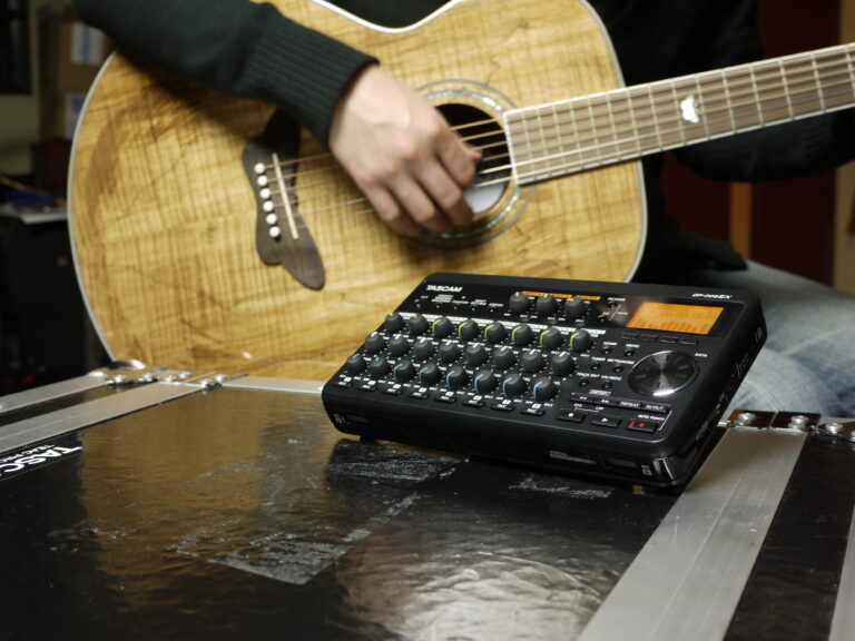 Le corps d’une guitare acoustique, avec une main jouant les cordes Au premier plan, un Tascam DP-008EX Pocketstudio