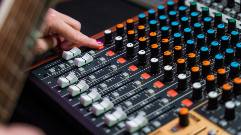 Angled view at a Tascam Model 12 mixing console with a musician pressing the record button on the first channel. A blurred guitar is partly visible on the left.